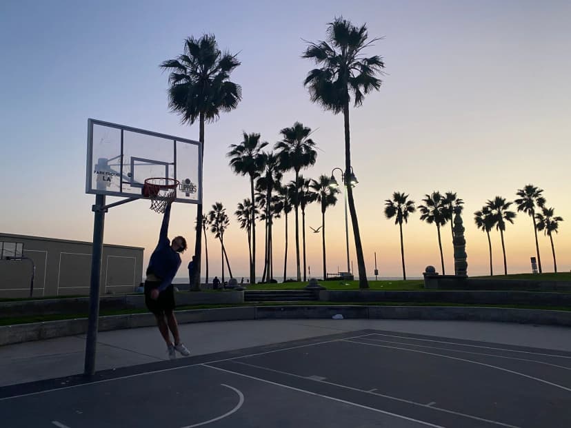 Joven jugando al baloncesto en una cancha al aire libre con palmeras al atardecer, ejemplo de deporte y tiempo libre durante un año escolar en USA.